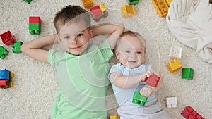 Portrait of baby and older boy lying on carpet next to colroful toys and bricks and looking up in camera. Concept of