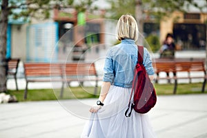 A portrait of the back of a girl holding marsala backpack wearing blue denim shirt and grey tulle skirt. A girl walks in