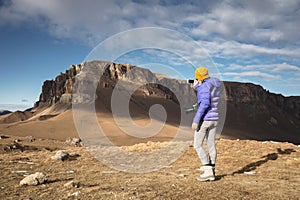 Portrait from the back of a traveling girl in a down jacket with a cap taking pictures of an epic landscape with rocks