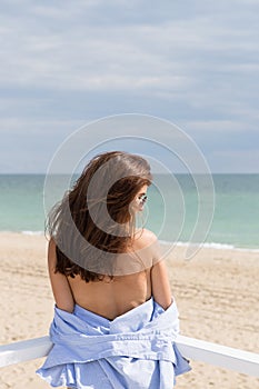 Portrait from the back of young girl standing on a background of beach, sand and sea. She wearing black swimwear, blue shir