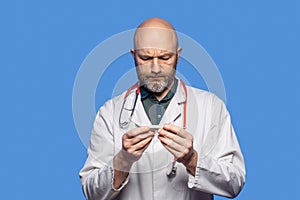 Portrait of bald doctor on blue background. Male health care worker in 40s, with grey beard holding white digital thermometer,