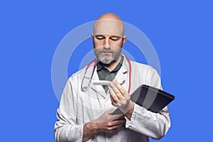 Portrait of bald doctor on blue background. Male health care worker in 40s, with grey beard holding white digital thermometer,