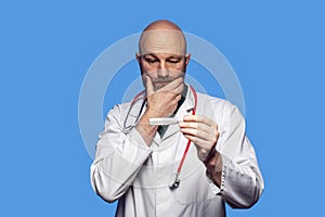 Portrait of bald doctor on blue background. Male health care worker in 40s, with grey beard holding white digital thermometer,