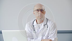 Portrait bald male doctor nodding head sitting on table front laptop in medical office. Practitioner doctor listening
