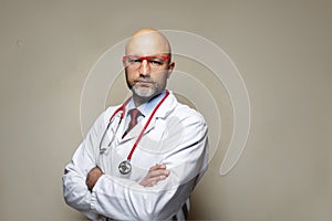 Portrait of a bald male doctor wearing white uniform and red stethoscope and glasses and blue shirt and red tie. Light cream
