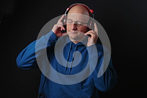 Portrait of a bald man listening to music on headphone. Male in his 40s, wears blue hoodie and glasses. Dark background. Eyes