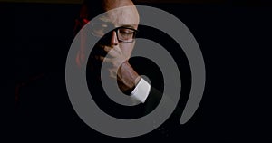 Portrait of a bald sad man in glasses and a business suit with a tie, it is on a black background. He`s loading his gun