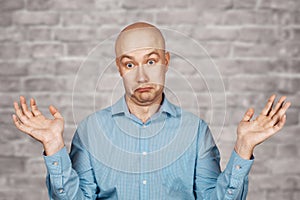 Portrait bald unsure man doesn`t understand anything and spreads his hands. guy in a blue shirt on white brick wall background in