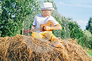 Portrait of barefoot boy in hat on haystack in field. Playing small guitar, ukulele. Light sunny day. Side view. Outdoor