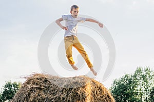 Portrait of barefoot boy in white short and yellow trousers jumping on haystack in field. Outdoor activity. Side view