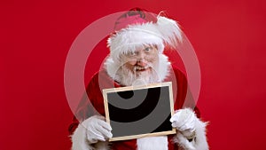 Portrait Beard Santa Claus in Red Suit Looks Into Camera and Peeks Out From Behind Black Sign in His Hands With