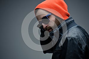 Portrait of a bearded brutal man in sunglasses and orange hat and black leather jacket, isolated on a grey background.