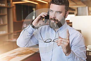 Portrait of bearded businessman in shirt,holding glasses in hand,lifting his index finger up, and talking on cell phone.