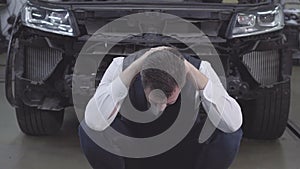 Portrait of bearded caucasian man sitting in front of broken car and holding hands together. Desperate businessman at