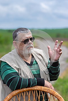 Portrait of bearded Caucasian senior man in black sunglasses leant on a wicker chair