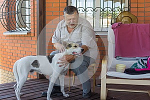 Portrait of bearded Caucasian senior man stroking his white mixed breed dog while sitting on veranda