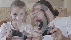 Portrait of a bearded dad and cute little daughter playing video games on gaming console sitting on the couch at home