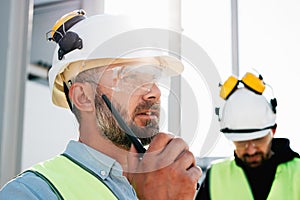 Portrait of bearded engineer wearing protect glasses and helmet use walkie-talkie on construction site. Team of builders control