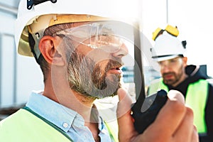 Portrait of bearded engineer wearing protect glasses and helmet use walkie-talkie on construction site. Team of builders control