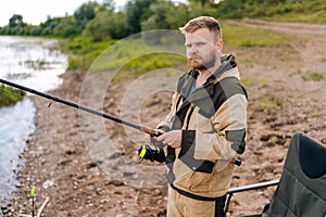 Portrait of bearded fisher man holding spinning casting rod standing on bank waiting for bites on water river at summer