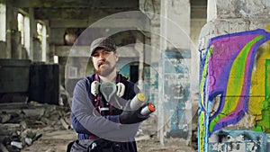 Portrait of bearded guy graffiti painter standing in old empty building with graffiti painting in background and holding