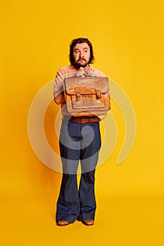 Portrait of bearded hairy man, shy scientist nerd in flared jeans standing with briefcase over yellow background. Retro