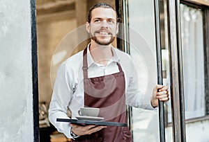 Portrait of bearded happy barista man working waitress in cafe open entrance door and serving drinks on a tray in front of the