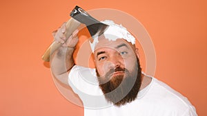 Portrait of bearded hipster man shaving his head with an ax axe, hatchet looking to the camera