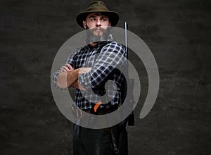 Portrait of a bearded hunter in a fleece shirt and hat standing with a rifle behind his back.