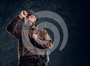 Portrait of a bearded hunter with rifle holding hand on hat and looking sideways. Studio photo against a dark wall