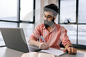 Portrait of bearded Indian businessman in glasses using laptop analyzing working project results, writing notes siting