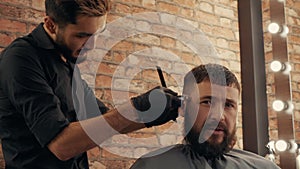 Portrait bearded man getting haircut in barbershop. Hairstylist cutting hair with electric razor in male salon. Close up