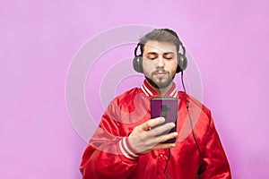 Portrait of a bearded man in the headphones standing against the background of a pink wall with a smartphone in his hand, looking