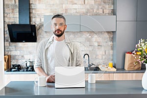 portrait of bearded man standing at counter with laptop and cups of coffee and looking at camera