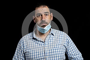 Portrait of bearded man in a white and blue plaid shirt wearing a wrongly fitted surgical mask to protect himself from coronavirus