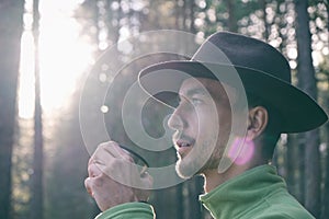 Portrait of bearded millennial man in felt hat drinking hot tea in woods on sunset. Tourist adventurer on trail in forest. Local