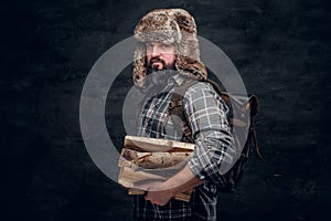 Portrait of a bearded woodcutter with a backpack dressed in a plaid shirt and trapper hat holding firewood. photo