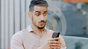 Portrait bearded young amazed handsome business man stands in city looking in mobile phone with shocked expression on