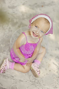 Portrait of beautiful 3 years old girl in pink summer dress and pink kerchief. Child sits on ground and looks in camera. View of