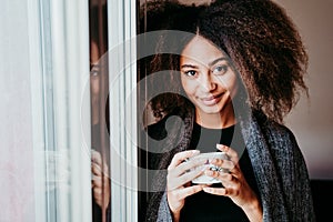 portrait of beautiful afro american young woman by the window holding a cup of coffee. Lifestyle indoors