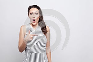 Portrait of beautiful amazed young brunette woman in striped dress standing and looking at camera with surprised face and pointing