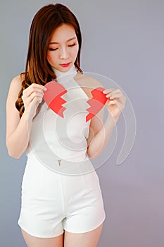 Portrait beautiful asia woman on gray background, unhappy in valentine day concept, model is holding red half heart in hand, broke