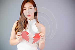 Portrait beautiful asia woman on gray background, unhappy in valentine day concept, model is holding red half heart in hand, broke