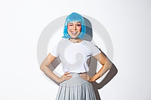 Portrait of beautiful asian girl in blue party wig showing tongue and winking silly, standing over white background in