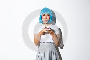 Portrait of beautiful asian girl in blue wig licking lips as holding delicious cake, standing over white background