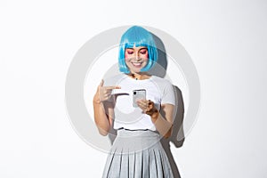 Portrait of beautiful asian girl pointing finger at smartphone, smiling pleased, standing in blue short wig and