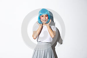 Portrait of beautiful asian girl in schoolgirl costume and blue wig, smiling kawaii at camera, standing over white