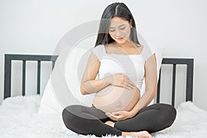Portrait of beautiful Asian pregnant woman sit on bed and touch and look to her belly also smiling. Concept of good take care baby