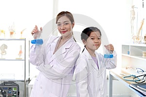 Portrait of beautiful Asian science teacher and adorable schoolgirl in lab coat holding blue chemical flasks and standing back to