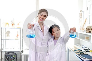Portrait of beautiful Asian science teacher and adorable schoolgirl in lab coat standing and showing blue chemical flasks